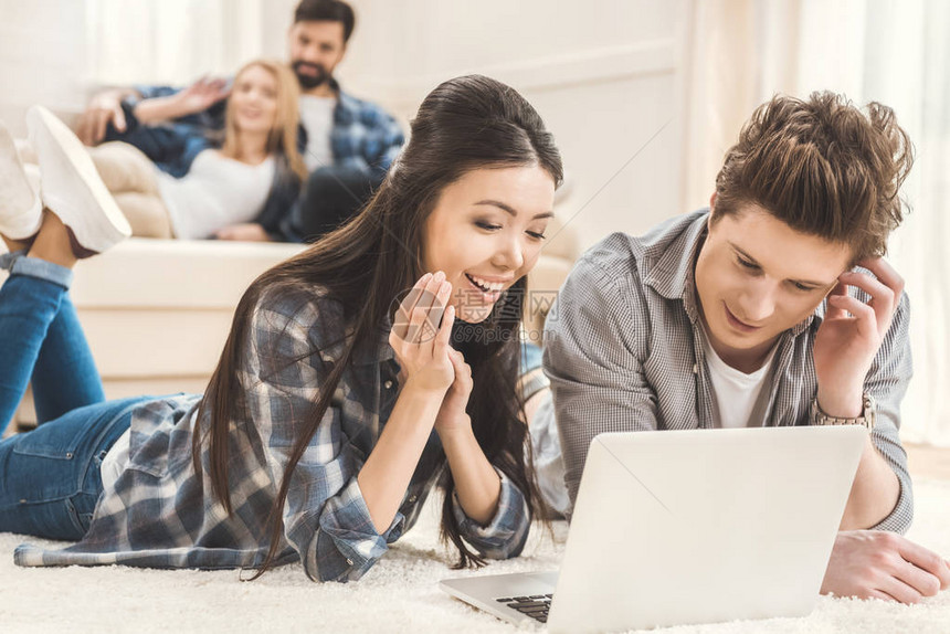 坐在地毯上并使用笔记本电脑与另一对夫妇一起坐在沙发上的背景面部的年图片