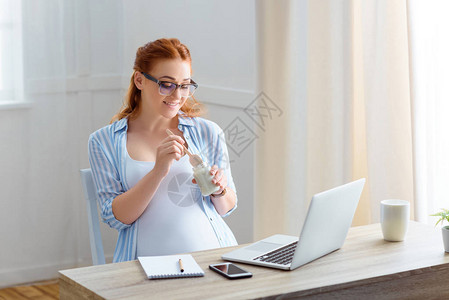 微笑的红发孕妇坐在桌边吃酸奶图片