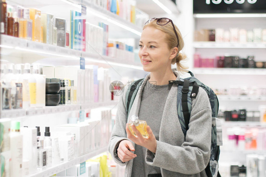 金发碧眼的年轻女旅行者穿着外套和旅行背包在机场免税店选择香水休闲女士在美容店随时地测试图片