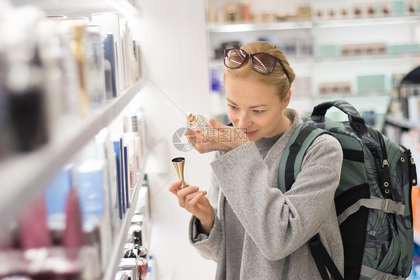 金发碧眼的年轻女旅行者穿着外套和旅行背包在机场免税店选择香水休闲女士在美容店随时地测试图片