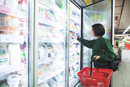 一个提着篮子的女孩从超市的冰箱里拿冷冻食品女孩在商店里挑选商品图片