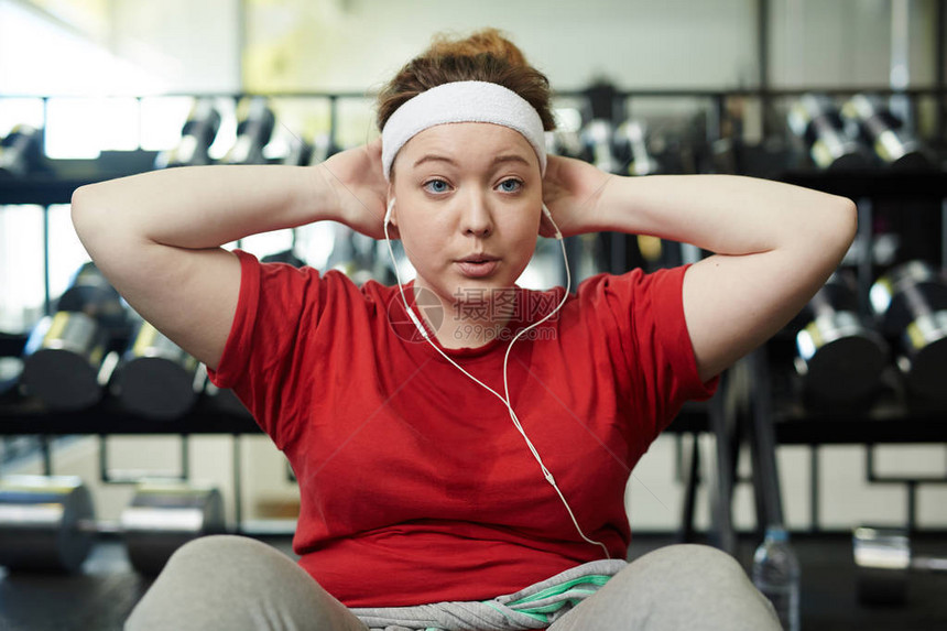 年轻肥胖女在健身房锻炼时做仰卧起坐锻炼努力呼吸并用耳机图片