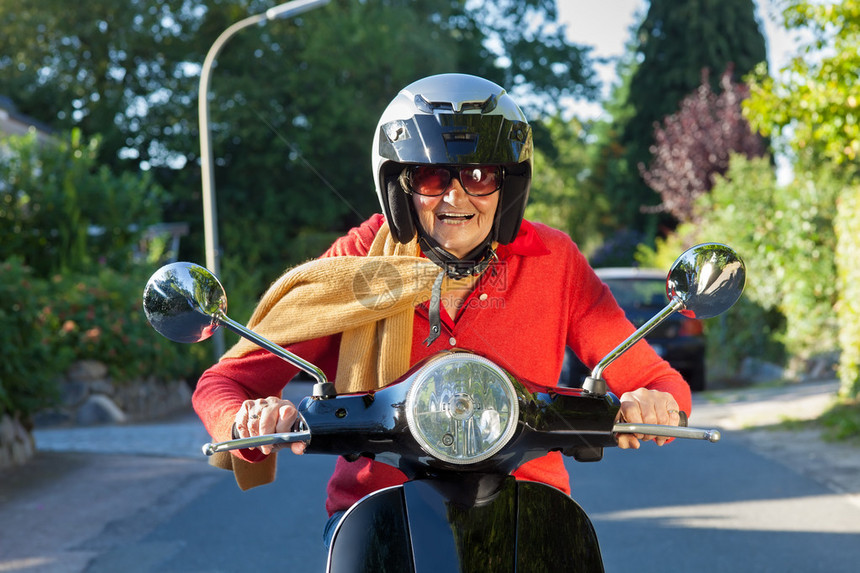 戴着头盔太阳镜和围巾的高级女士直接对着镜头骑着摩托车图片