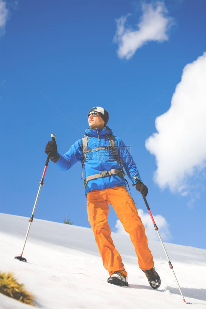 一个背着登山杖和背包的年轻人在蓝天和云彩的映衬下在山上的雪地上行走图片