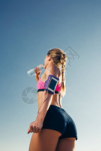 使用智能手机在蓝天上操作臂带箱式饮用水的耳机运动女运动员低角图片