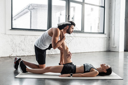 在健身房帮助年轻运动女运动员在瑜伽垫上伸腿图片