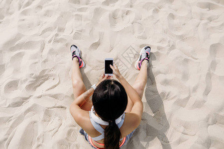 手持智能手机和耳机坐在沙滩上的女图片