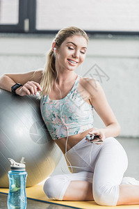 戴着耳机的快乐女运动员在健身房的健身球附近用智能手机图片