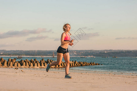 运动女运动员用耳机和智能手机在沙滩上与海后面跑步时使用手持臂图片