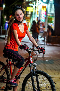 城市夜骑自行车戴自行车头盔的女孩夜生活和城市背景中的路人发光的窗户和图片
