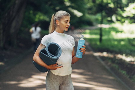健身女人在清晨公园准备瑜伽穿着时尚的运动装蓝色瓶装水和瑜伽垫健图片