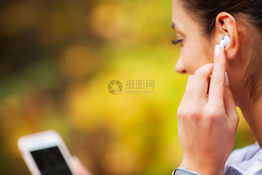 20岁服触摸蓝牙耳膜手持机的女子在绿色公园休息时图片