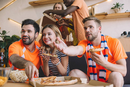 年轻男子用手指着和朋友吃披萨在家看足球比赛图片