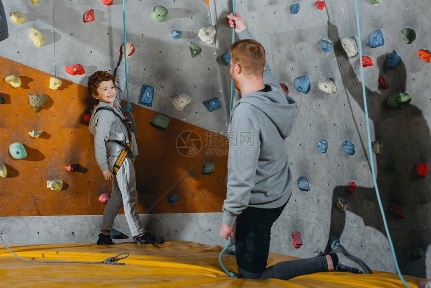 小男孩在攀爬绳子上站在体育馆的墙壁旁边与他父图片
