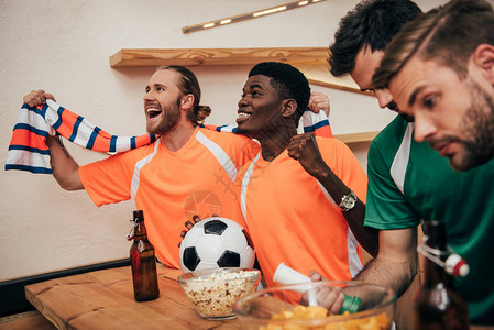 身穿橙色T恤和围巾的多元文化足球迷微笑着用球庆祝胜利图片