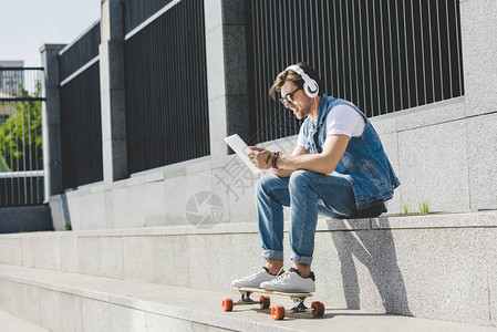 有滑板的时尚青年男子在街上用耳机和平板图片