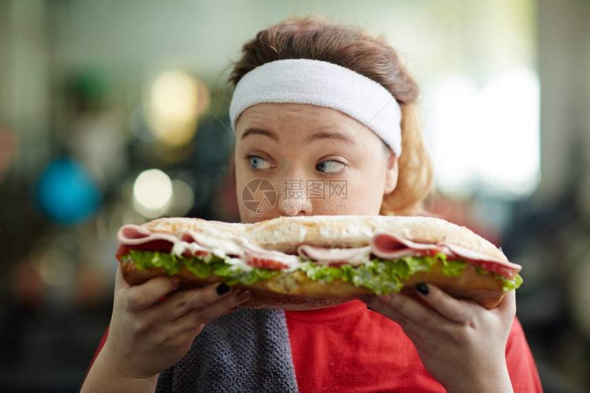 可爱超重女人的近视肖像在她面前拿着大胖子三明治秘密吃着它仔细地环顾四周裹图片