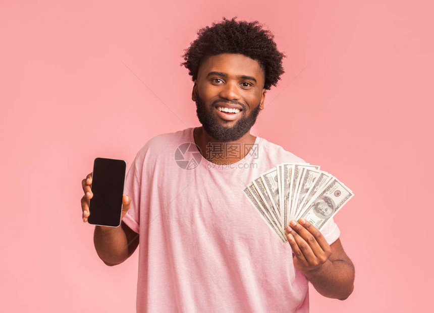 通过电话赚钱快乐的非洲男人展示空白智能手机屏幕和一大笔美元在粉红背景上图片