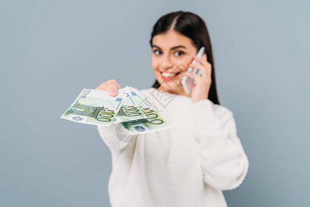 在白毛衣上给欧元钞票和谈论在灰色上隔离的智能手机时以微笑的漂亮女孩图片