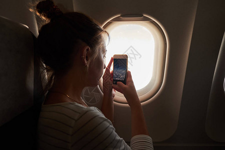一个年轻的女孩看着飞机的窗外乘客从飞机窗外看女孩用手机拍图片