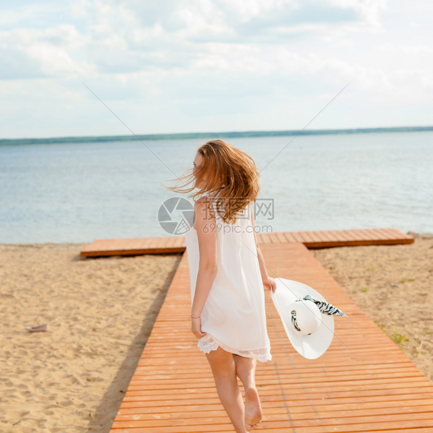 穿着白帽子白色裙子的浪漫女孩转向海滩上长尾木头码喜悦和幸福的概念图片