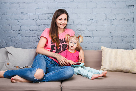 美丽的年轻母亲使用智能手机与坐在房间里沙发上的一个两岁女儿玩耍图片