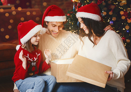 可爱的小女孩打开礼物盒和年轻父母们一起在圣塔帽在圣诞节晚夜图片