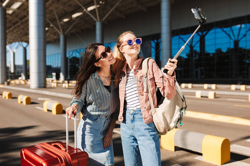 两个戴着墨镜的漂亮女孩在机场附近的户外肩上带着手提箱和背包图片