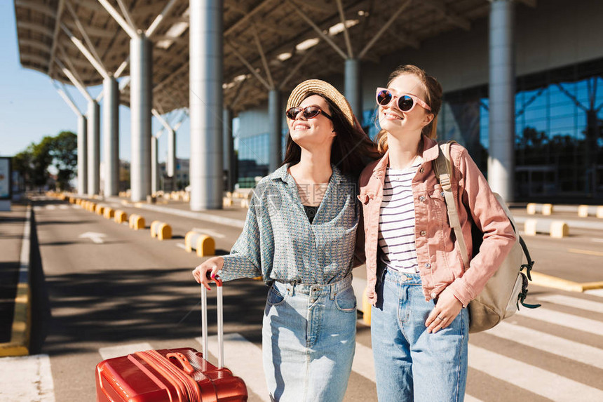 两个戴着太阳镜的漂亮微笑女孩带着红色手提箱和背包在机场附近的户外欢图片