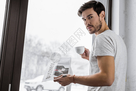 英俊的年轻人拿着一杯咖啡和报纸看着镜头图片