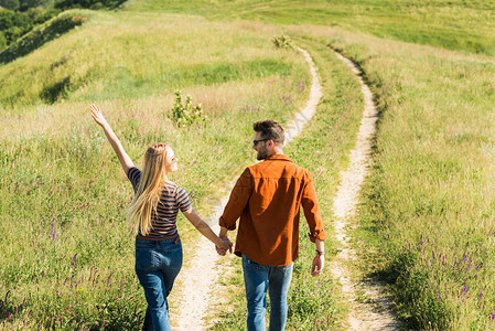 年轻夫妇手牵穿过乡村草地的后视图图片