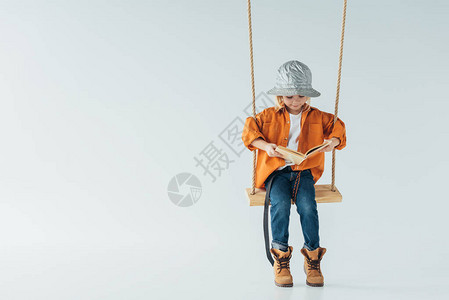 穿着牛仔裤和橙色衬衫的可爱孩子坐在摇摆和阅读书图片