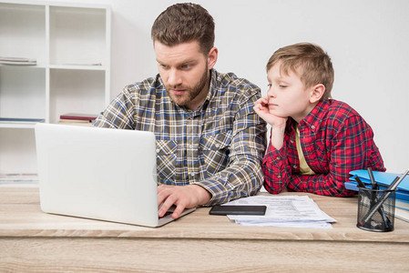 在家庭办公室与儿子一起使用笔记本电脑的商图片