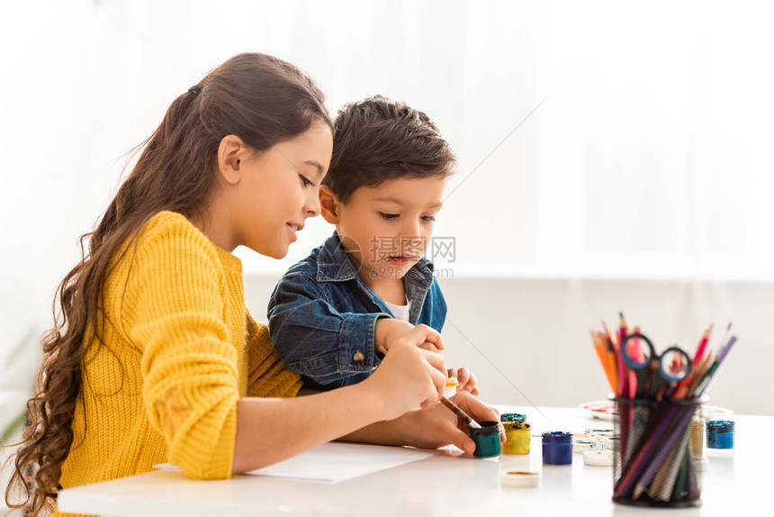 专注的兄妹坐在桌旁一起用颜料画图片