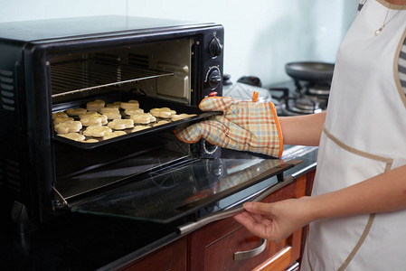 身着烤炉手套的不可辨认妇女将未经加工的饼干烘烤板放在家用厨房背景图片