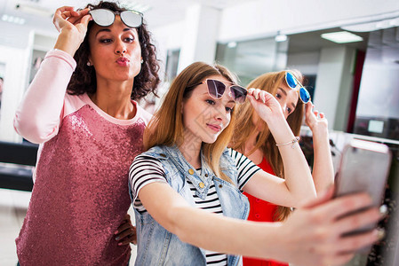 三个年轻时髦的女朋友在购物中心举起时尚墨镜带着智能手机自拍图片