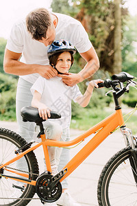 父亲把头盔戴在儿子身上而男孩站在自行车旁看远图片