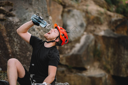 穿着头盔的英俊年轻旅行者在岩石前面用塑料瓶装塑图片