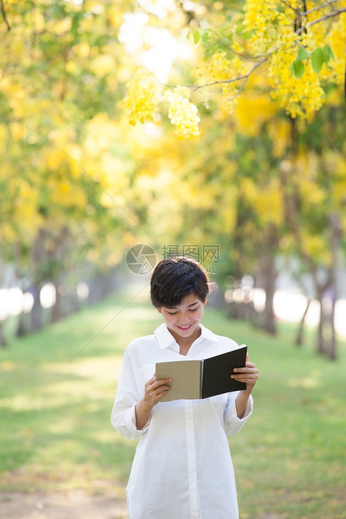 年轻美丽的亚洲女人的肖像站在黄色花朵盛开的公园中夏天季和阅读一本充满图片