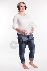 怀孕的金发女郎穿着白色t恤和牛仔裤在白色背景上听耳机上的音图片