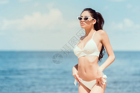 美丽的年轻美女穿着太阳镜和白泳衣在度假胜地靠近海背景图片