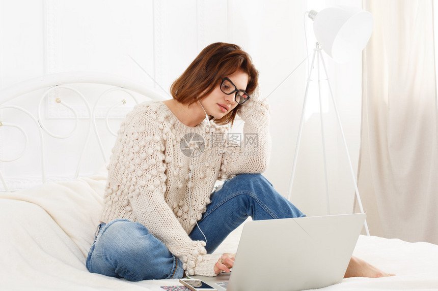 年轻女子在床上从笔记本电脑上看书眼镜和耳机的全神贯注的女孩与手机有魅力的女人穿着便服在白色的床上互联网自由职业者高调图片