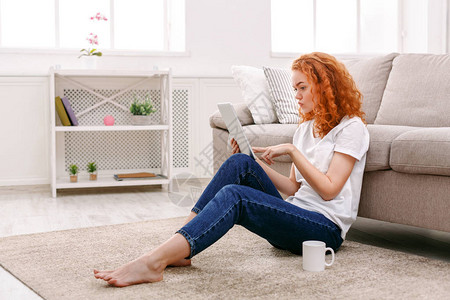 年轻体贴的红发女孩在平板电脑上阅读休闲的年轻女子坐在背景图片
