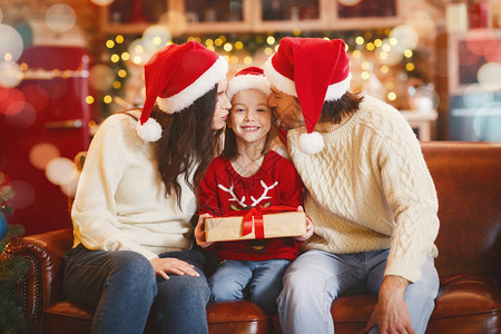 带着圣诞礼物的快乐小女孩被父母亲用圣塔帽吻过而不是装饰背景图片
