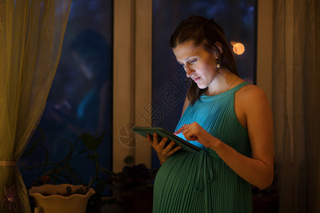 穿着蓝裙子的孕妇晚上在窗户旁边使用家里的平板药片图片