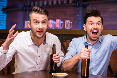 在酒吧看电视两个快乐的年轻人坐在酒吧里喝着啤图片