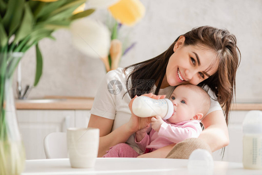 护理婴儿微笑的年轻女子用瓶中的配方喂养新生儿女人轻地拥抱她的孩图片
