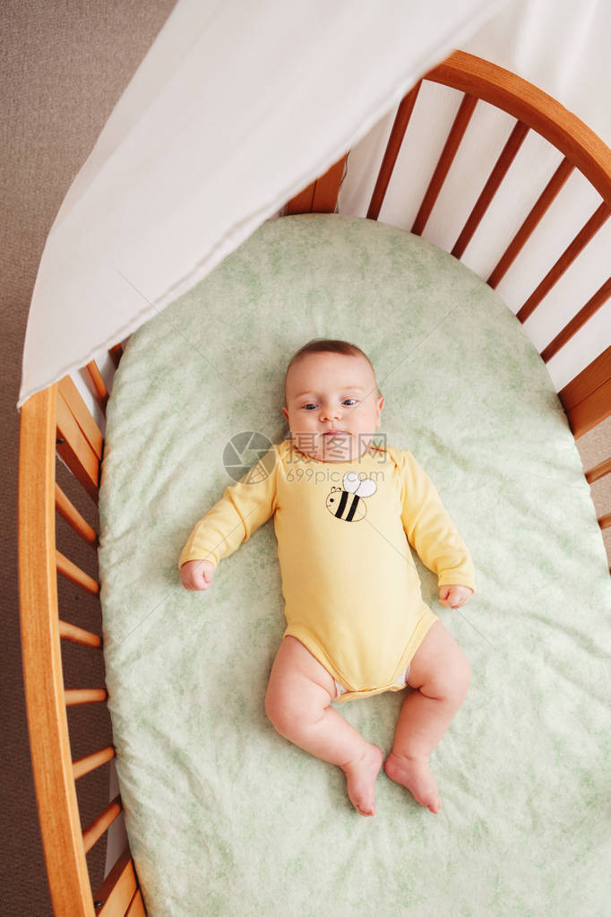 身穿黄色连体衣独自躺在婴儿床靠近窗边的可爱风趣可爱白人小婴儿的画像图片