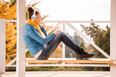 坐在白色耳机和用智能手机监听音乐的木板凳上的年轻美女的侧面肖图片