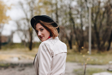 穿着战壕大衣和帽子的年轻微笑着的女士在秋天公园散图片